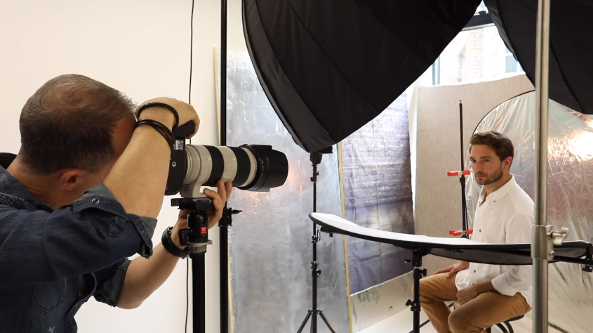 A model is sitting in a studio being photographed by a professional photographer.